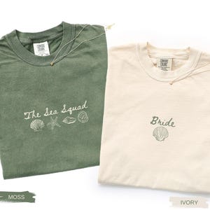 May include: A moss green t-shirt with the text The Sea Squad and four shell illustrations, and an ivory t-shirt with the text Bride and a single shell illustration. Both shirts feature a crew neck and a Comfort Colors brand label at the collar.