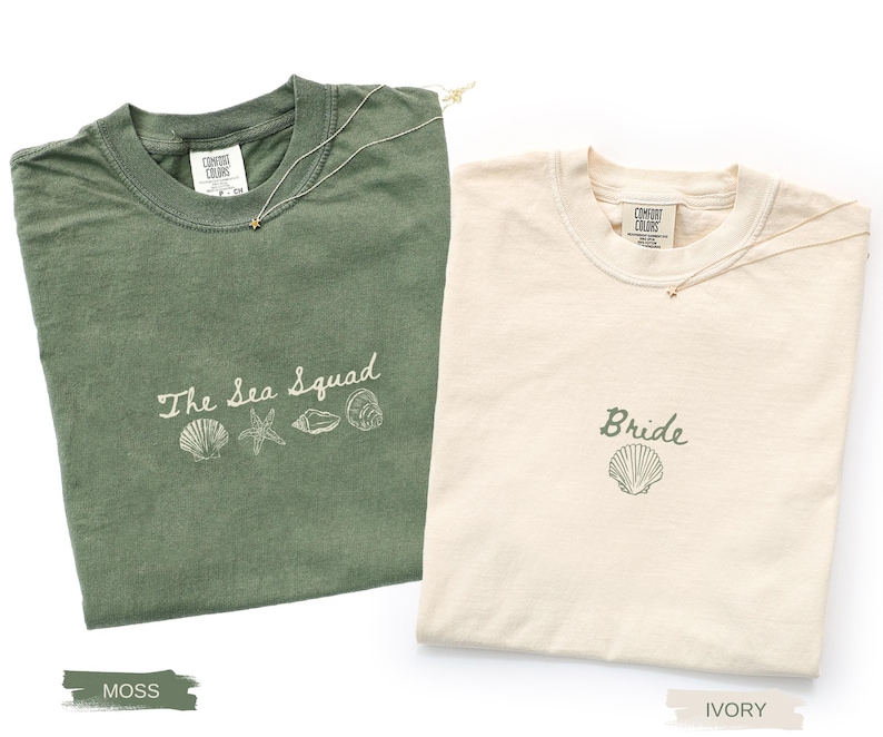 May include: A moss green t-shirt with the text The Sea Squad and four shell illustrations, and an ivory t-shirt with the text Bride and a single shell illustration. Both shirts feature a crew neck and a Comfort Colors brand label at the collar.