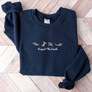 Peut inclure: Un sweat-shirt bleu marine avec un graphique blanc de cinq requins et le texte "Respect The Locals".