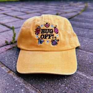 May include: Mustard yellow baseball cap with the embroidered text "BUG OFF!" surrounded by colorful bug and flower designs. The cap has a curved brim and a classic six-panel design, perfect for outdoor activities.