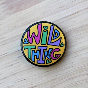 May include: A round button with a yellow background and black outline. The words "WILD THING" are written in colorful letters with pink, green, purple, and blue.