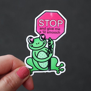 May include: A green frog with pink hearts for eyes holds a pink stop sign that says "STOP and give me a lil smooch".