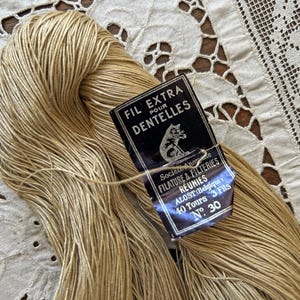 May include: A skein of light beige thread with a black rectangular paper label attached. The label features a cat illustration and text reading FIL EXTRA POUR DENTELLES, SOCIETE ANONYME FILATURE & FILTERIES REUNIES ALOST BELGIQUE 40 TOURS 3 FILS N 30.