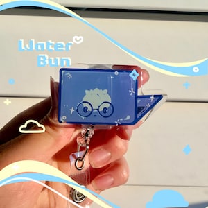 Puede incluir: Un portatarjetas de acrílico azul y transparente con el texto "Water Bun" y un personaje de dibujos animados con gafas. El portatarjetas tiene un cordón retrácttil y un clip metálico.