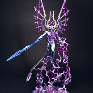 May include: A miniature figurine of a humanoid figure with a metallic body, adorned with purple and blue lightning effects. The figure holds a blue spear and has large, wing-like structures. The base is a textured, circular platform.