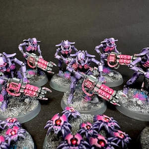 Può includere: Una collezione di miniature, con guerrieri robotici viola e rosa con grandi armi. Le figure sono su basi grigie, con verniciatura dettagliata e un'estetica fantascientifica. Sono presenti anche creature più piccole simili a ragni.