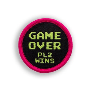 May include: A pink and black circular patch with the text "GAME OVER" in green pixelated letters and "PL2 WINS" in smaller green pixelated letters.
