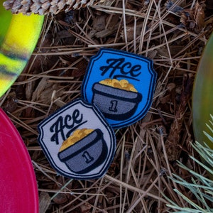 May include: Two embroidered patches with the word "Ace" and a cartoon image of a pot with a number "1" on it. The patches are on a brown background of pine needles.