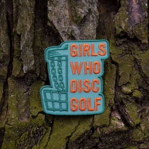 May include: A teal blue embroidered patch with an orange outline. The patch features a disc golf basket and the text "Girls Who Disc Golf".
