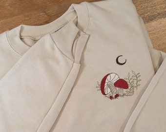 Pode incluir: Camisola bege com design bordado de cogumelos vermelhos, uma lua crescente e folhagem. A parte superior de manga comprida tem gola redonda e punhos canelados. O desenho está na parte superior esquerda.