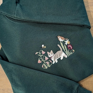 Può includere: Felpa verde scuro con ricamo. Il design presenta un gatto grigio e bianco, un uccello, un'ape e fiori rosa. La felpa è piegata su una superficie di legno.