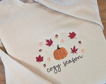 May include: Beige sweatshirt with an embroidered design featuring a pumpkin, fall leaves, and the words "cozy season" in a script font. The design is in shades of orange, red, and brown.