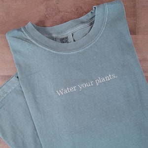 Puede incluir: Camiseta verde salvia con la frase "Water your plants" bordada en blanco. La camiseta está doblada sobre una superficie de madera. El nombre de la marca es visible en el cuello.