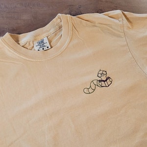 Op de afbeelding: Een lichtgele longsleeve t-shirt met een geborduurd ontwerp van een worm met een bril die een boek leest. Het shirt heeft een ronde hals en een relaxte pasvorm. Het label luidt "Soft Colors" en de maat is M-M.