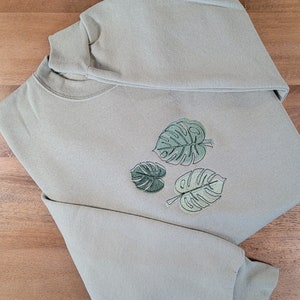 Puede incluir: Una sudadera verde salvia con tres hojas de monstera bordadas en diferentes tonos de verde. La parte superior de manga larga está doblada, mostrando el cuello redondo y el diseño de hojas en la parte delantera. La tela parece suave y cómoda.
