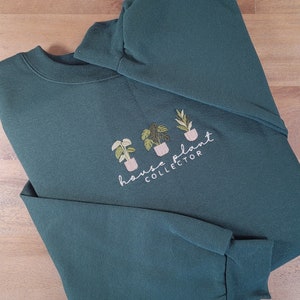 Puede incluir: Una sudadera verde oscuro con el texto bordado "house plant collector" y tres pequeñas ilustraciones de plantas en macetas. La parte superior de manga larga está doblada sobre una superficie de madera.