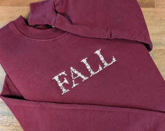 Puede incluir: Una sudadera burdeos con la palabra "FALL" bordada en blanco, con una fuente floral. Las mangas largas están dobladas y la sudadera está sobre una superficie de madera.