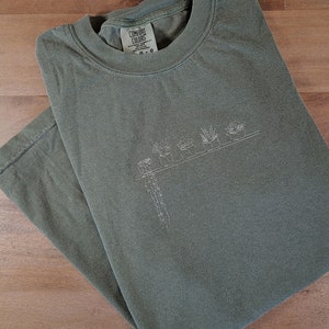 Puede incluir: Camiseta verde oliva con un dibujo lineal blanco de plantas en macetas. La camiseta está doblada sobre una superficie de madera. La etiqueta dice "Comfort Colors". El diseño presenta una fila de suculentas y cactus.