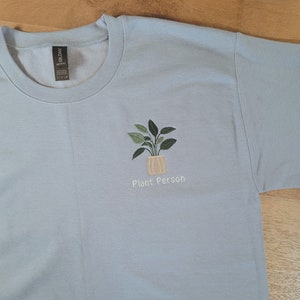 Puede incluir: Sudadera azul claro con un diseño de planta bordada en una maceta beige. El texto "Plant Person" está bordado debajo de la planta. La sudadera está hecha de un material suave.
