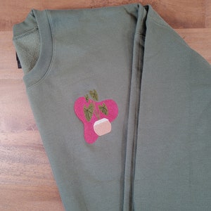 Puede incluir: Sudadera verde oliva con un diseño de planta bordada en rosa y verde. La planta está en una maceta blanca. La sudadera está hecha de un material suave y tiene cuello redondo.
