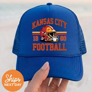 May include: A blue trucker hat with the words "KANSAS CITY FOOTBALL" in orange lettering. The hat features a red football helmet graphic with the numbers "1960". The hat is held by a hand.