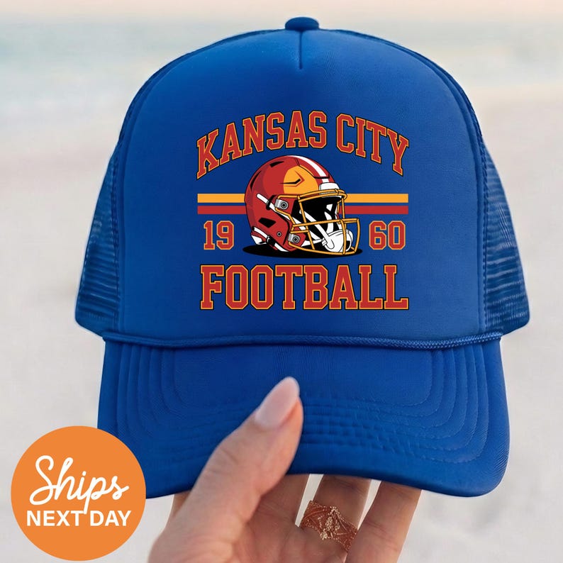 May include: A blue trucker hat with the words "KANSAS CITY FOOTBALL" in orange lettering. The hat features a red football helmet graphic with the numbers "1960". The hat is held by a hand.
