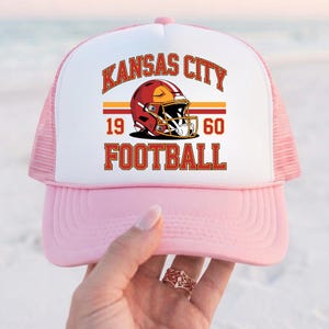 May include: Pink and white trucker hat with a graphic featuring a football helmet and the words "Kansas City Football 1960". The hat has a pink brim and mesh back.