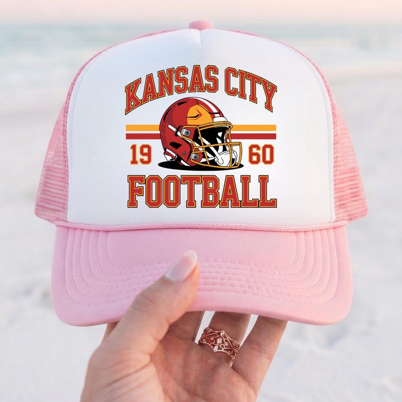 May include: Pink and white trucker hat with a graphic featuring a football helmet and the words "Kansas City Football 1960". The hat has a pink brim and mesh back.