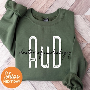May include: An olive green sweatshirt with the white letters "AuD" and the words "doctor of audiology" in a cursive font. The sweatshirt has a crew neck and long sleeves. An orange circle with the words "Ships NEXT DAY" is in the lower left corner.