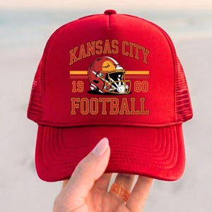 May include: Red trucker hat with the text "Kansas City Football" and a football helmet graphic. The hat includes the years "1960" and has a mesh back.