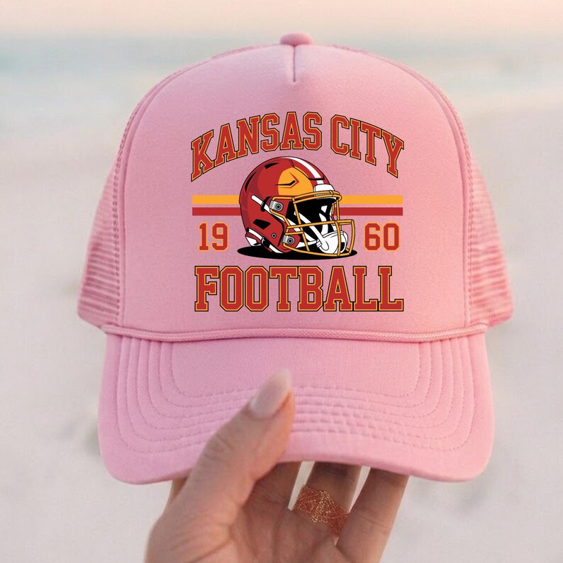 May include: Pale pink trucker hat with the words "Kansas City Football" in orange and brown. The hat features a red and gold football helmet graphic, with the numbers "19" and "60" below. The back of the hat is mesh.