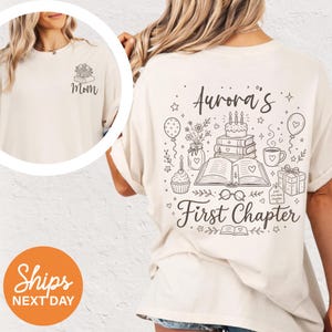 May include: Cream-colored t-shirt with a black graphic design. The front features the word "Mom" with a floral and book design. The back displays the text "Aurora's First Chapter" with illustrations of a cake, books, and balloons.