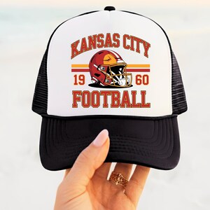 May include: Black and white trucker cap with a white front panel displaying "Kansas City Football" and a football helmet graphic. The cap includes the year "1960" in red. It has a curved peak and a mesh back.