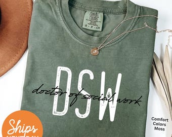 May include: An olive green t-shirt with the text "DSW doctor of social work" in white. The shirt has a crew neck and short sleeves. A gold necklace with a pendant is draped on the shirt. The shirt is labeled "Comfort Colors Moss".