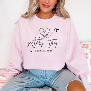 a woman sitting on a bed wearing a pink sweatshirt