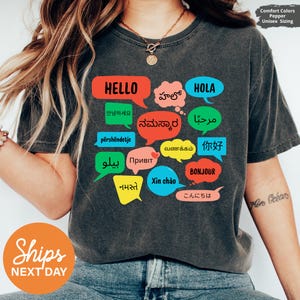 May include: Dark gray unisex t-shirt with colorful speech bubbles displaying the word "Hello" in multiple languages. The shirt also features the words "Hola", "Bonjour", and "Xin chào". The shirt is labeled "Comfort Colors" and "Unisex Sizing".