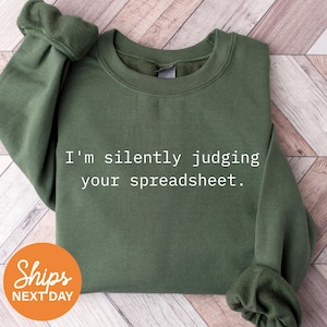 May include: Olive green sweatshirt with the text "I'm silently judging your spreadsheet." The sweatshirt has a crew neck and long sleeves. An orange circle with the words "Ships Next Day" is in the lower left corner.