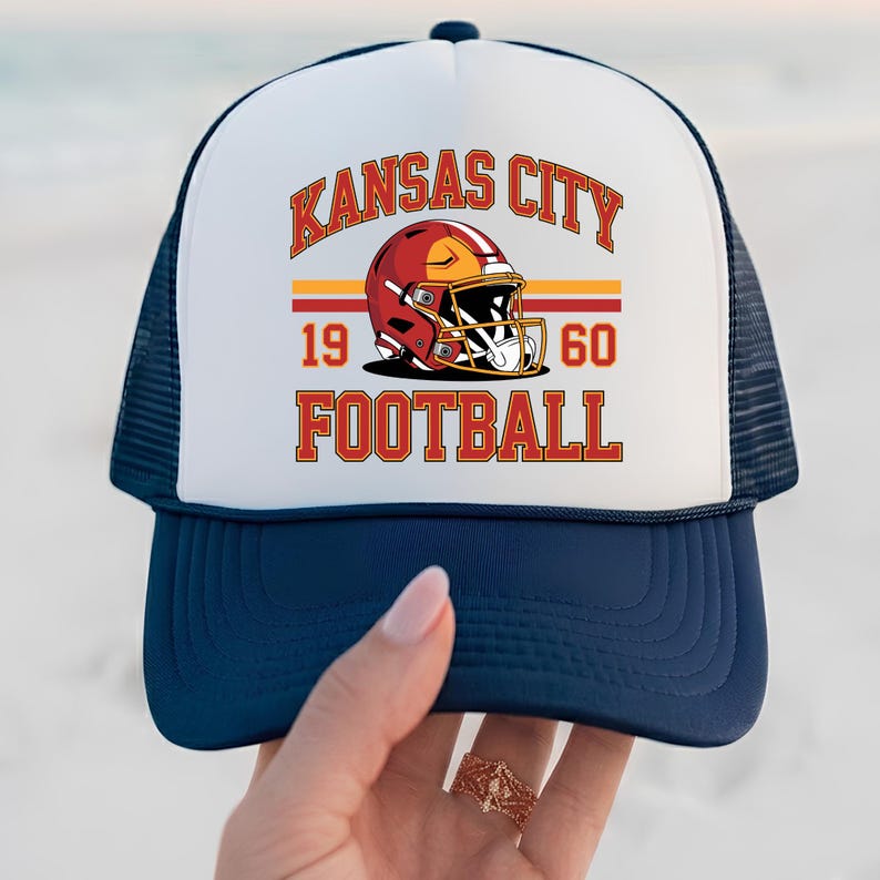 May include: A navy blue and white trucker hat with the text "KANSAS CITY FOOTBALL" in orange lettering. The hat features a football helmet graphic and the numbers "1960". The hat has a mesh back.