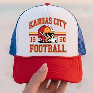 May include: A red, white, and blue trucker hat with the text "Kansas City Football" and a football helmet graphic. The hat has a red peak, a white front panel, and a blue mesh back. The text "1960" is also displayed.