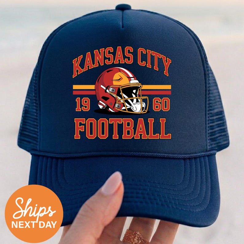 May include: A navy blue trucker cap with a mesh back. The front displays the words "Kansas City Football" in orange, a football helmet graphic, and the numbers "1960".
