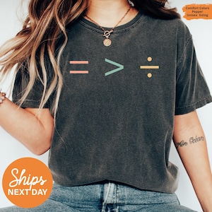 May include: Dark gray unisex t-shirt with the text "= > ÷" in coral, teal, and yellow. The shirt has a crew neck and short sleeves. The words "Comfort Colors, Pepper, Unisex Sizing" are printed on the top right.