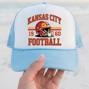 May include: A light blue and white trucker hat with the text "KANSAS CITY FOOTBALL" in red and gold. The hat features a football helmet graphic and the numbers "1960". The hat has a mesh back.