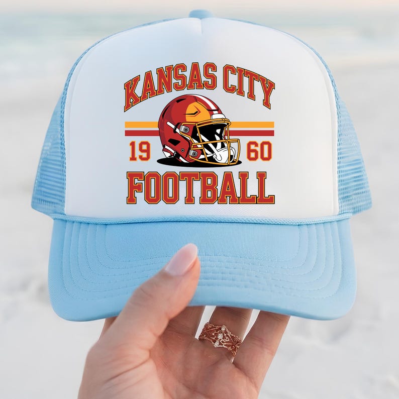 May include: A light blue and white trucker hat with the text "KANSAS CITY FOOTBALL" in red and gold. The hat features a football helmet graphic and the numbers "1960". The hat has a mesh back.