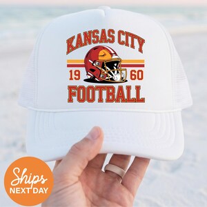May include: White trucker hat with the words "Kansas City Football" in red and gold. The hat has a mesh back and a curved peak. The design includes a football helmet and the numbers "1960".