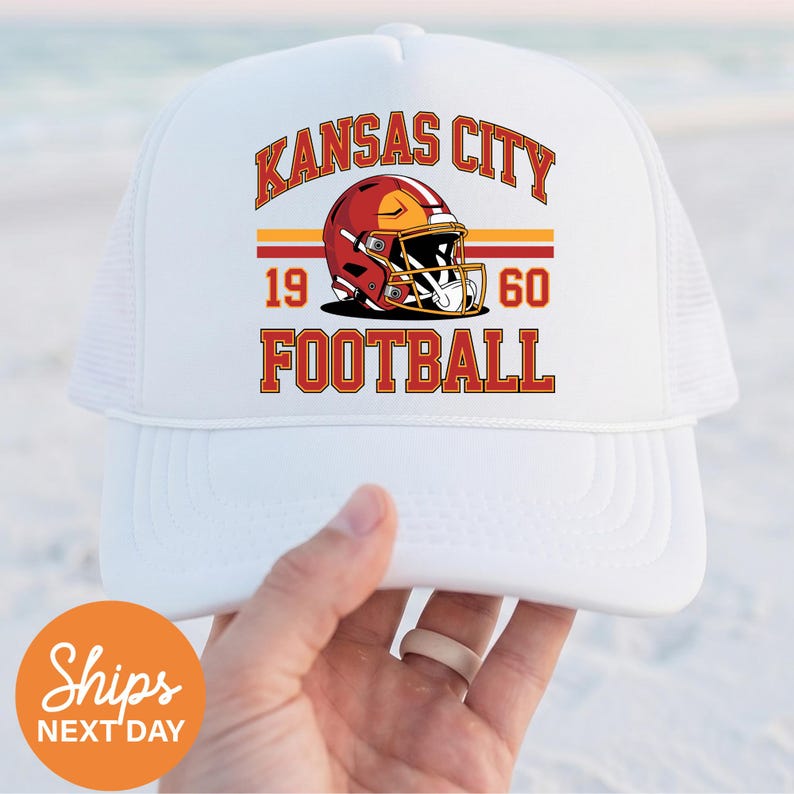 May include: White trucker hat with the words "Kansas City Football" in red and gold. The hat has a mesh back and a curved peak. The design includes a football helmet and the numbers "1960".