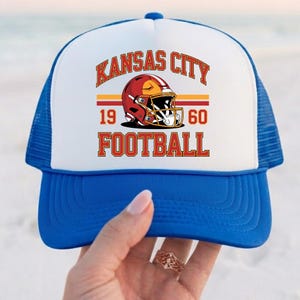May include: A blue and white trucker hat with the text "KANSAS CITY FOOTBALL" in red and gold. The hat features a football helmet graphic and the numbers "1960". The hat has a blue brim and mesh backing.