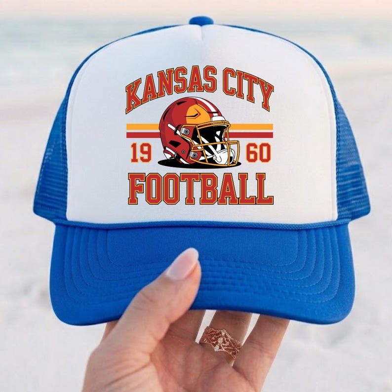 May include: A blue and white trucker hat with the text "KANSAS CITY FOOTBALL" in red and gold. The hat features a football helmet graphic and the numbers "1960". The hat has a blue brim and mesh backing.