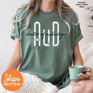 May include: Moss green t-shirt with the letters "AuD" in white, above the words "doctor of audiology" in a script font. The shirt is a comfort colors t-shirt.