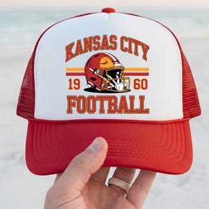 May include: Red and white trucker hat with the text "KANSAS CITY FOOTBALL" and a football helmet graphic. The hat has a red peak and mesh back, with the text "1960" below the helmet.