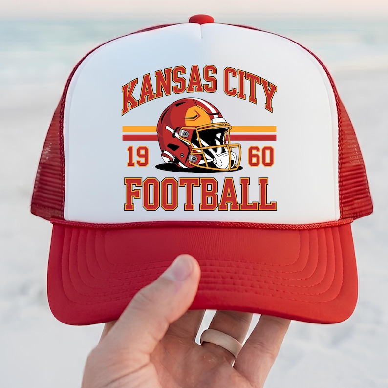 May include: Red and white trucker hat with the text "KANSAS CITY FOOTBALL" and a football helmet graphic. The hat has a red peak and mesh back, with the text "1960" below the helmet.